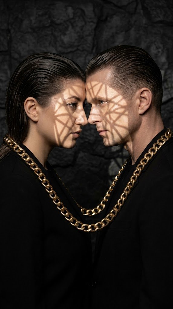 A professional symmetrical photo of the man and the woman from the uploaded images. They are facing each other, their foreheads almost touching. A thick golden metal chain is draped over their shoulders, binding them together in a perfect loop. Both have perfectly slicked-back hair and minimal wet-look makeup. The background is a dark textured stone. A sharp light beam (gobo effect) creates a geometric pattern of light across their faces, highlighting the golden chain's reflection on their skin. Sculptural poses, total black and gold color palette, high-fashion edito