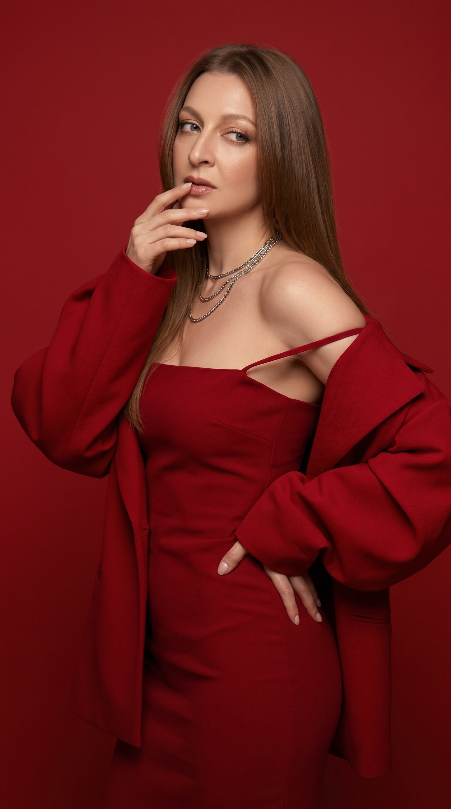 Ультрареалистичная glamorous monochrome red/editorial съёмка в минималистичной студии. Девушка стоит в динамичной cinematic pose, как будто резко оборачивается назад через плечо, чтобы посмотреть в сторону. Корпус развёрнут в три четверти, талия скручена, спина мягко выгнута. Голова повёрнута назад, взгляд направлен направо из-под ресниц, soft mysterious expression. Одна рука поднята к лицу, пальцы слегка касаются губ и подбородка, вторая рука лежит на талии с изогнутым запястьем.
На ней облегающее monochrome red dress с тонкими бретелями и straight neckline. Одна тонкая бретелька небрежно спадает с плеча, создавая effortless sensual styling. Поверх платья oversized red jacket со спущенными плечами и exaggerated sculptural sleeves. Рукава очень объёмные, мягко драпируются вокруг рук, создавая couture silhouette. Вся одежда выполнена в одном насыщенном глубоком алом оттенке, seamless monochrome styling.
Волосы длинные, гладкие, блестящие, very dense blowout hair с мягким luxurious объёмом. Макияж glamorous: sculpted contouring, nude glossy lips с более тёмным lip contour, glowing highlight, soft blurred skin finish. Layered silver chain necklaces.
Фон полностью однотонный насыщенно-красный, minimal studio backdrop без деталей. Мягкий cinematic studio lighting, dreamy diffusion, subtle haze, glossy highlights, low contrast soft glamour retouching, rich deep red color grading, smooth luxury beauty campaign aesthetic. Атмосфера luxurious, feminine, bold, mysterious, high fashion glamour aesthetic. Съёмка как для Valentino / Mugler beauty campaign, Canon G7X photography, ultra photorealistic skin texture, shallow depth of field, clean composition, monochrome red palette, highly detailed, 8K masterpiece.
9:16
