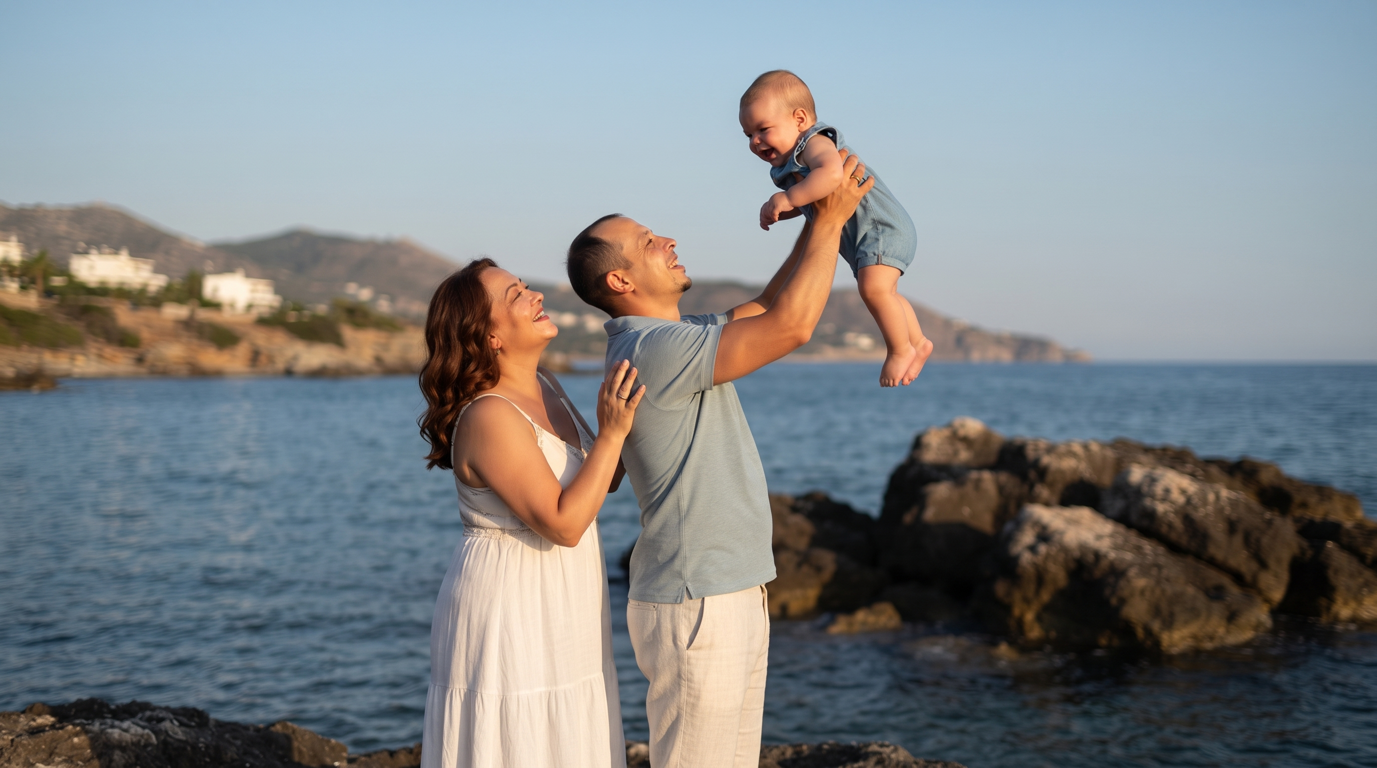 Create a photorealistic professional family photoshoot, using the attached images as strict references. Image 1 = the mother's identity (keep her exact face, round face shape, wavy chestnut-brown shoulder-length hair). Image 2 = the baby's identity (keep his exact face). Image 3 = the father's identity (keep his exact face, short dark hair with receding hairline, small goatee). Image 4 = composition, pose and clothing guide ONLY (do not copy the faces from it).

Composition exactly like image 4: the father stands in profile, joyfully lifting the baby high above his head with both arms fully extended, smiling up at him; the baby looks down at the father; the mother stands close behind the father, in profile, gazing up at the baby, both hands gently resting on the father's shoulder and back.

Clothing matching image 4: mother in a long white sleeveless sundress; father in a light grey-blue short-sleeved polo shirt and cream linen trousers; baby in a light blue denim romper, barefoot.

Setting: rocky seaside at golden hour, calm deep-blue sea, dark boulders, distant soft-focus hills with white houses, clear pale-blue sky. Warm low sunset sunlight, natural skin tones and texture, candid joyful mood, DSLR photo, 50mm lens, f/2.8, shallow depth of field. Aspect ratio 3:2.