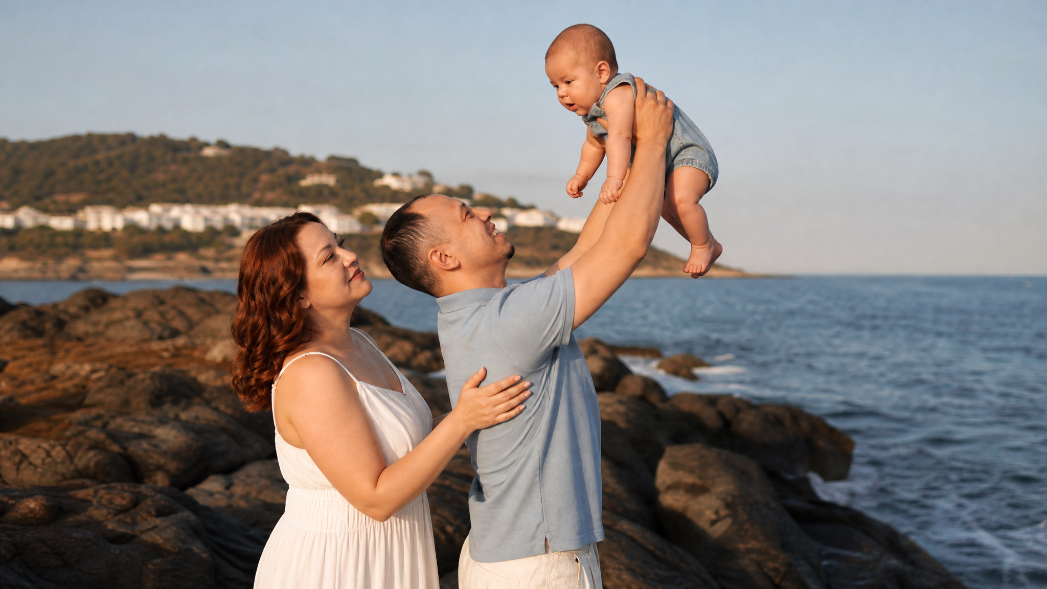 Create a photorealistic professional family photoshoot, using the attached images as strict references. Image 1 = the mother's identity (keep her exact face, round face shape, wavy chestnut-brown shoulder-length hair). Image 2 = the baby's identity (keep his exact face). Image 3 = the father's identity (keep his exact face, short dark hair with receding hairline, small goatee). Image 4 = composition, pose and clothing guide ONLY (do not copy the faces from it).

Composition exactly like image 4: the father stands in profile, joyfully lifting the baby high above his head with both arms fully extended, smiling up at him; the baby looks down at the father; the mother stands close behind the father, in profile, gazing up at the baby, both hands gently resting on the father's shoulder and back.

Clothing matching image 4: mother in a long white sleeveless sundress; father in a light grey-blue short-sleeved polo shirt and cream linen trousers; baby in a light blue denim romper, barefoot.

Setting: rocky seaside at golden hour, calm deep-blue sea, dark boulders, distant soft-focus hills with white houses, clear pale-blue sky. Warm low sunset sunlight, natural skin tones and texture, candid joyful mood, DSLR photo, 50mm lens, f/2.8, shallow depth of field. Aspect ratio 3:2.