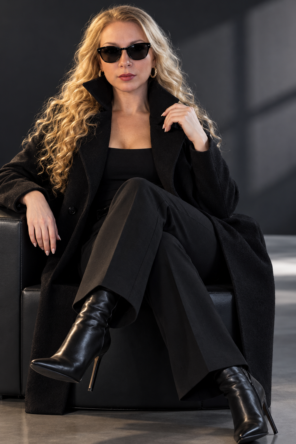 "Studio photo of a young, beautiful, stylish woman. She wears a black coat with the collar turned up, black sunglasses, long curly hair, tailored trousers, and high-heeled boots. She has bold, professional makeup. The scene is polished, high fashion, with a modern and chic aesthetic. Perfect lighting highlighting her features, capturing elegance and confidence."